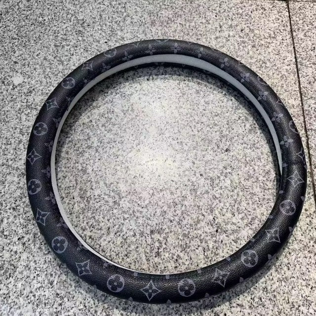 Black steering wheel cover with Louis Vuitton monogram pattern on a concrete surface